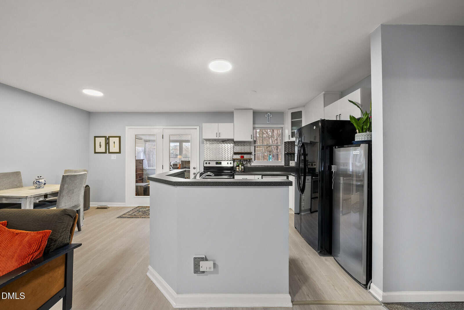Modern kitchen with light wood flooring, black appliances, white cabinets, and a breakfast bar. Dining area with a table and chairs is visible to the left, and glass doors lead outside in the background.
