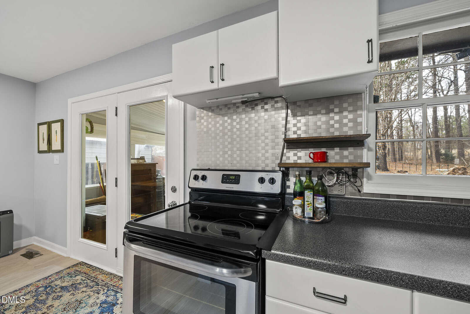 Modern kitchen with stainless steel stove, mosaic tile backsplash, black countertop, small bottles by the wall, a window showing trees, and glass doors leading to a sunroom with a piano and framed artwork.