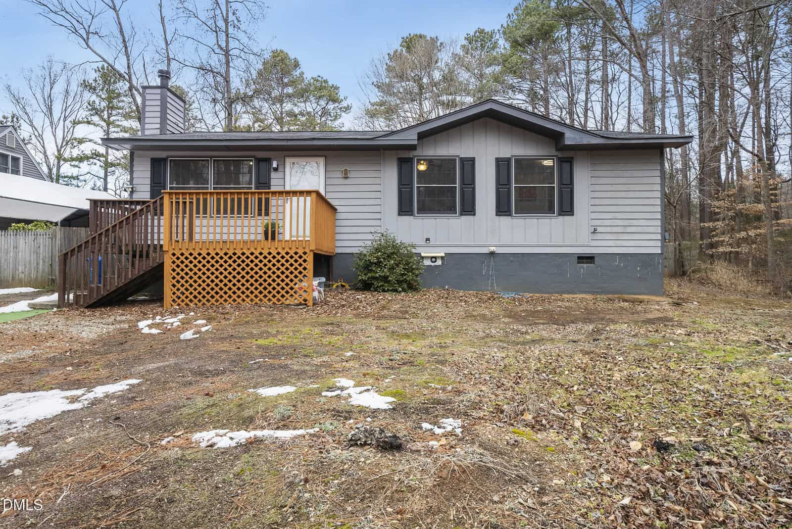 A single-story gray house with black shutters and a small wooden front porch sits on a patchy yard with sparse grass, some snow, and surrounded by trees under a clear sky.