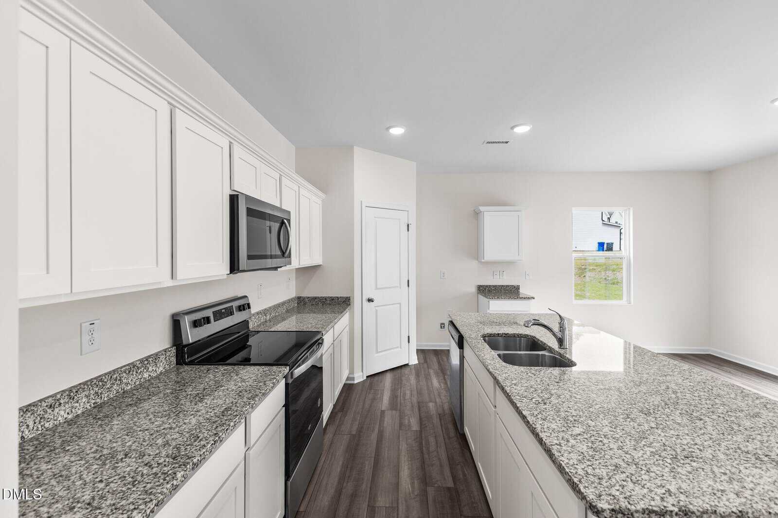 Modern kitchen with white cabinets, stainless steel appliances, granite countertops, a center island with a double sink, dark wood flooring, and a window letting in natural light.