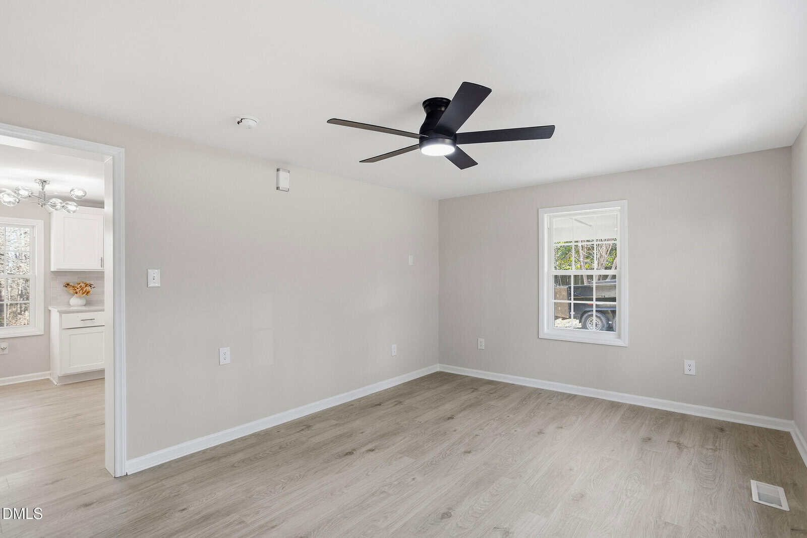 A bright, empty room with light wood flooring, beige walls, a large window, and a black ceiling fan. A doorway leads to a white kitchen area with cabinetry and a chandelier.