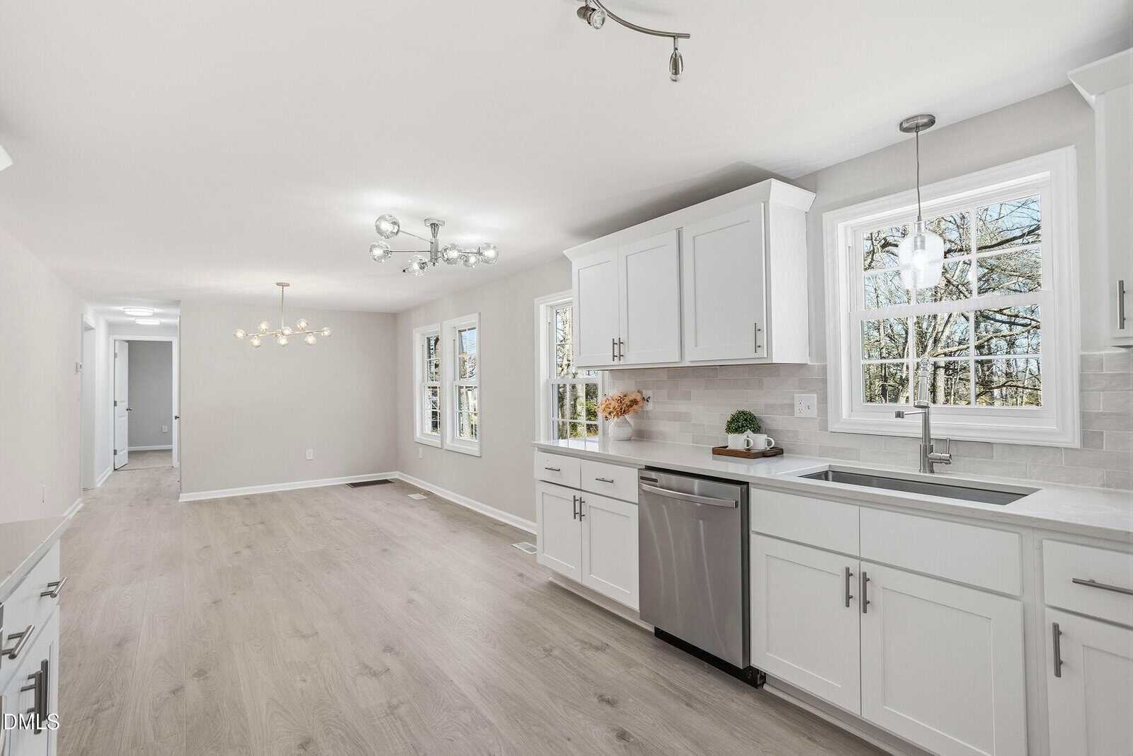 Bright, modern kitchen with white cabinets, stainless steel appliances, light wood floors, and large windows letting in natural light. Adjacent dining area features contemporary light fixtures and neutral walls.