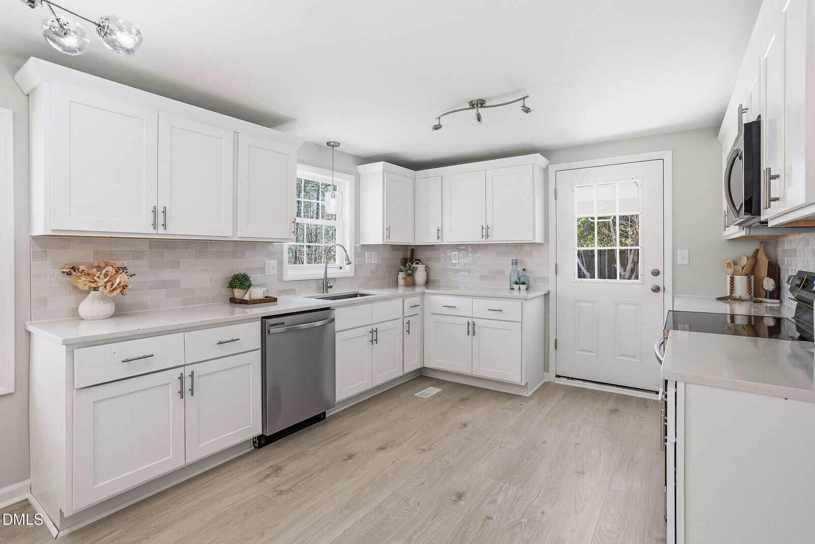 Bright, modern kitchen with white cabinets, stainless steel appliances, light wood flooring, tile backsplash, and a window and door letting in natural light. Minimalistic decor on countertops.