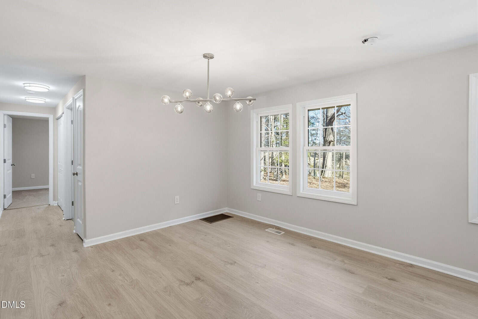 A bright, empty room with light wood flooring, pale gray walls, two large windows, and a modern chandelier. Sunlight streams in, and a hallway with several doors is visible on the left.