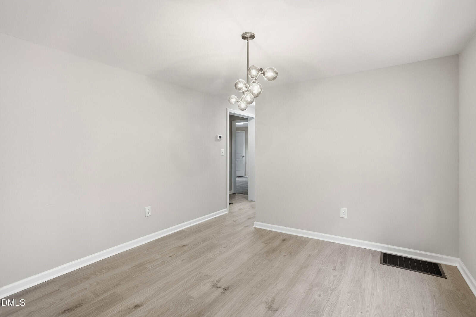 A bright, empty room with light wood flooring, plain white walls, a modern chandelier with globe lights, and an open doorway leading to a hallway.