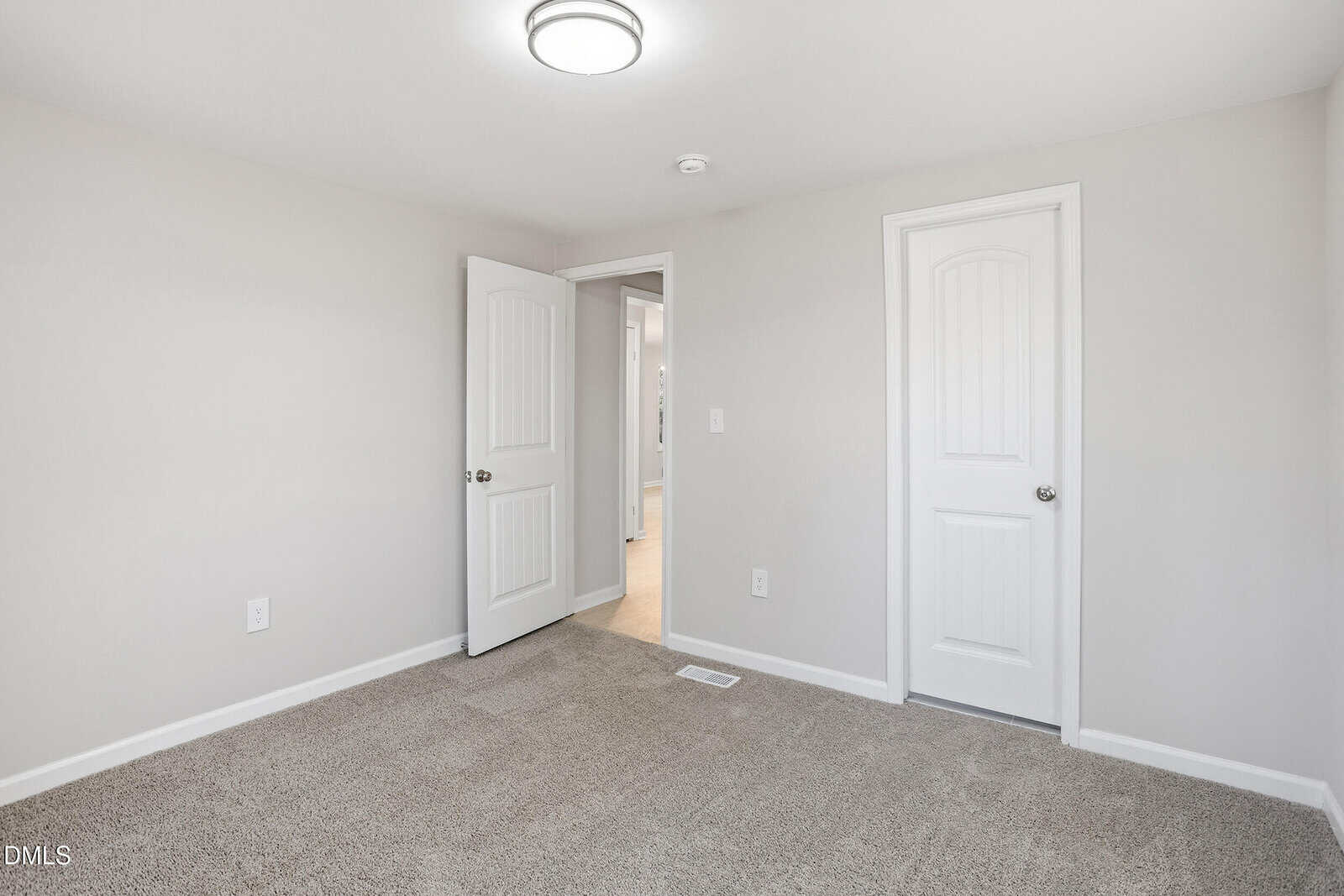 A small, empty room with beige carpet, light gray walls, a ceiling light, and two white doors—one open, showing a hallway with wood flooring, and one closed.