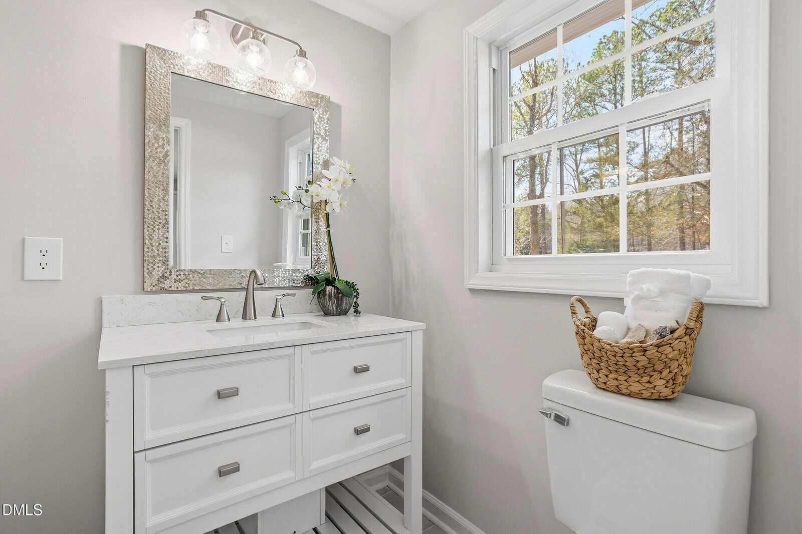 Bright bathroom with a white vanity, silver-framed mirror, potted orchid, and a window letting in natural light. A woven basket with towels sits on the toilet tank beside the vanity.