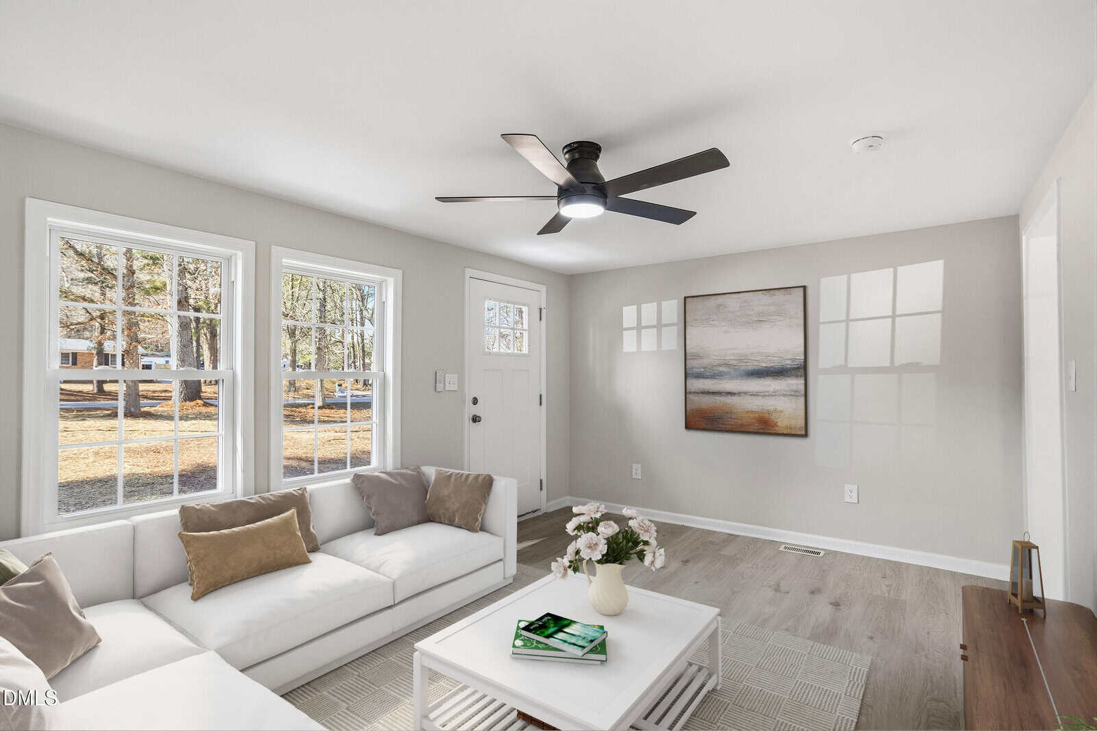 Bright living room with large windows, a white sectional sofa with cushions, a modern ceiling fan, a coffee table with flowers and books, light wood flooring, and a neutral abstract painting on the wall.