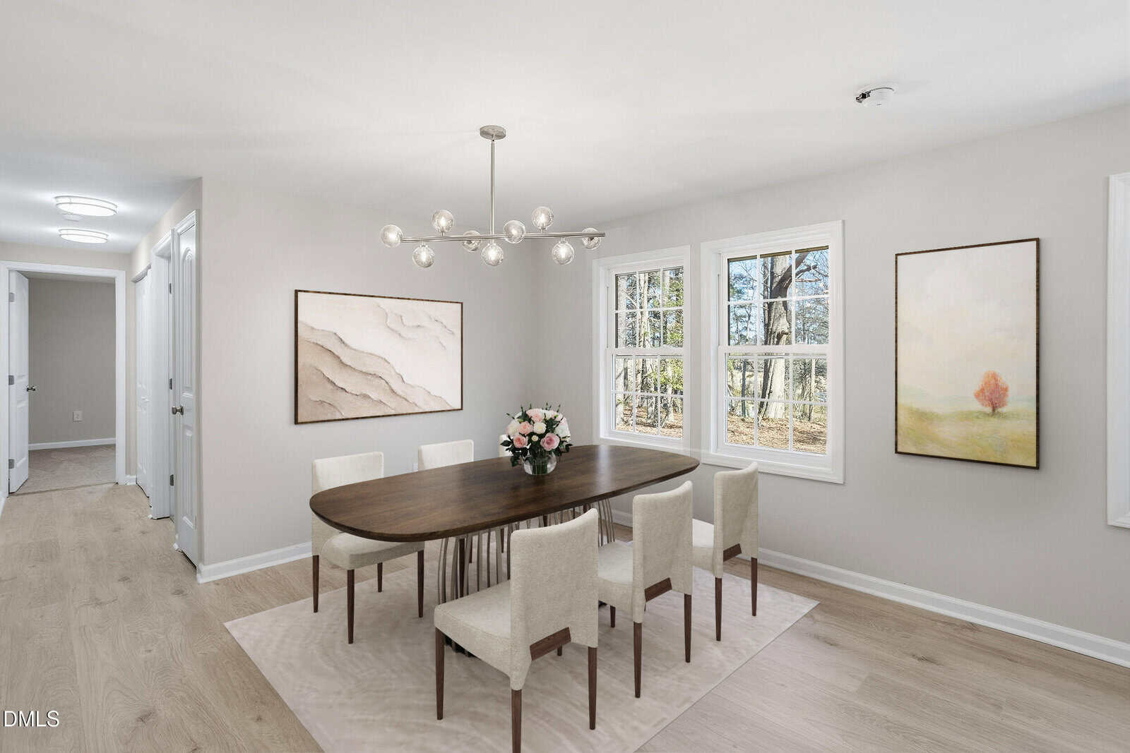 A modern dining room with a dark wooden table, six cream-colored chairs, a vase of flowers, light wood floors, neutral walls, large windows, contemporary chandelier, and two abstract paintings on the walls.
