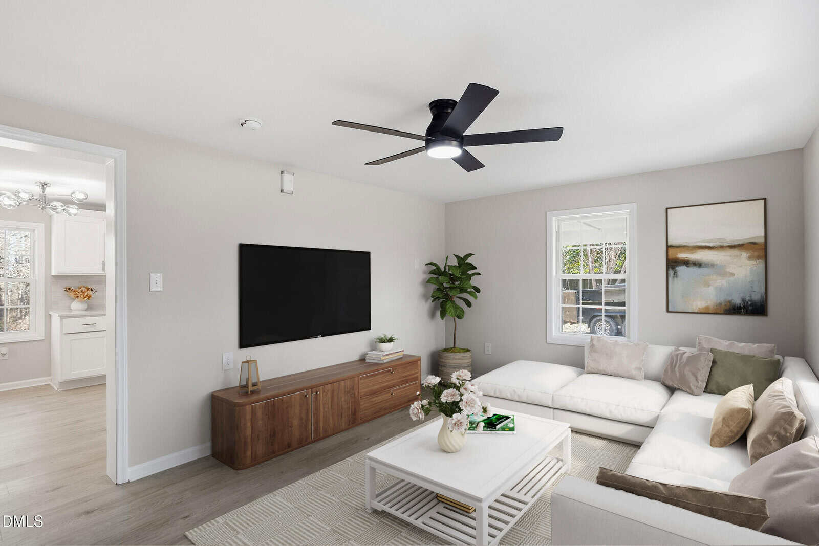 A modern living room with a white sectional sofa, wooden TV console, wall-mounted TV, coffee table with decor, large plant, abstract wall art, and a ceiling fan. A bright kitchen is visible through a doorway.