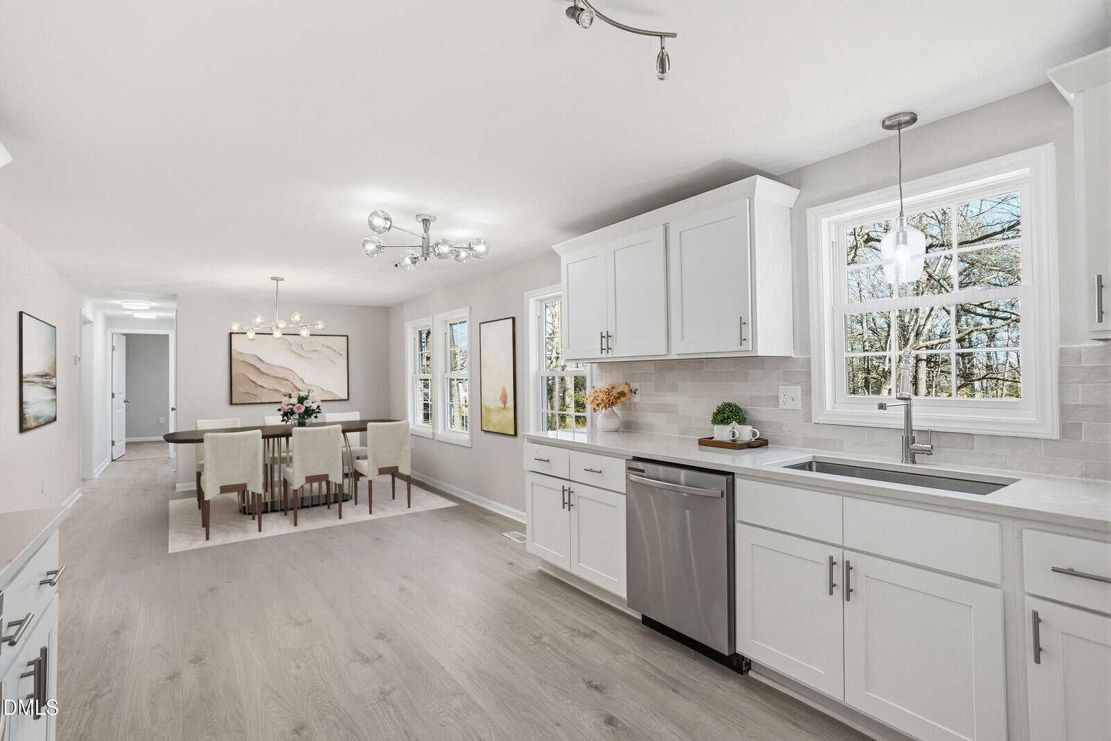 Modern kitchen with white cabinets, silver appliances, and large windows. Adjacent dining area features a table with six beige chairs, a flower centerpiece, and contemporary light fixtures. Bright, airy space with light wood flooring.