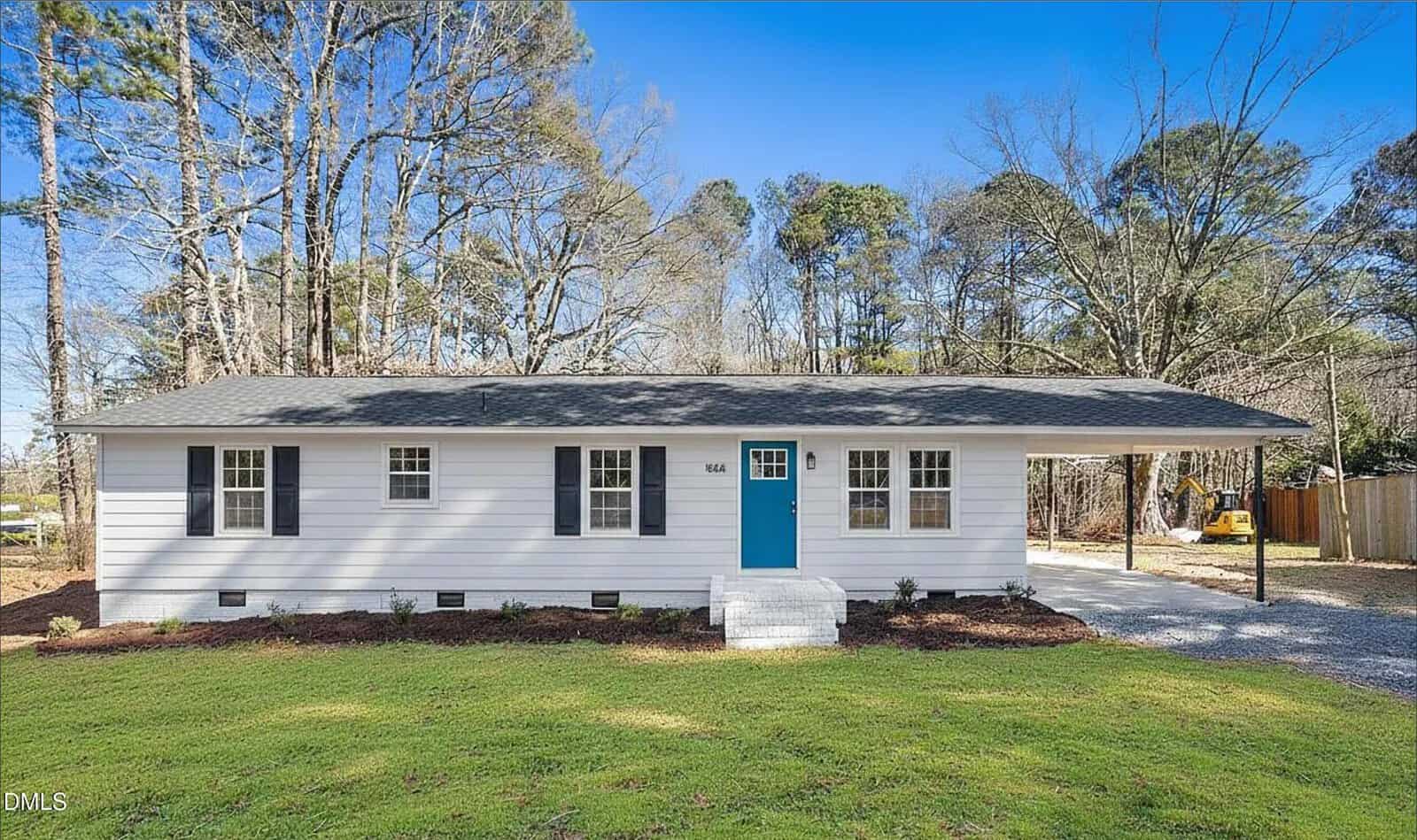 A single-story white house with dark shutters and a bright blue front door, set on a grassy lawn with trees in the background and a driveway leading to a carport on the right.