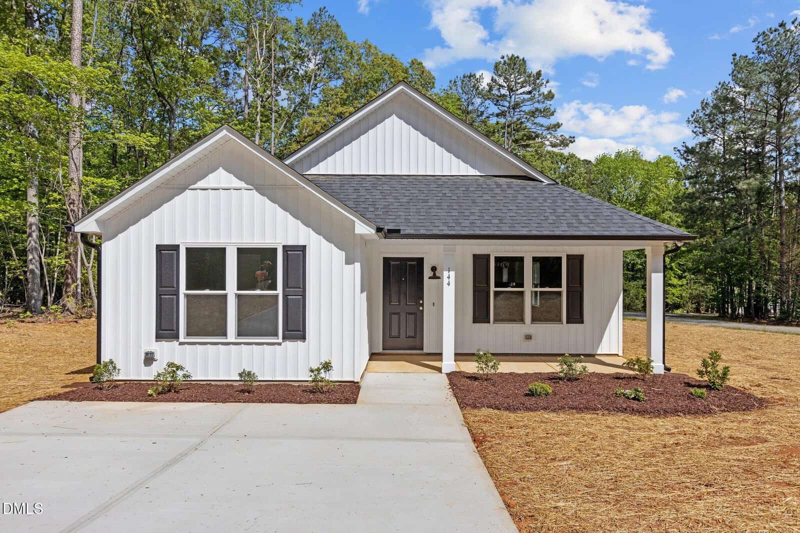 A white single-story house with black shutters and a covered front porch, set on a newly landscaped yard with fresh mulch and young shrubs, surrounded by tall trees under a partly cloudy sky.
