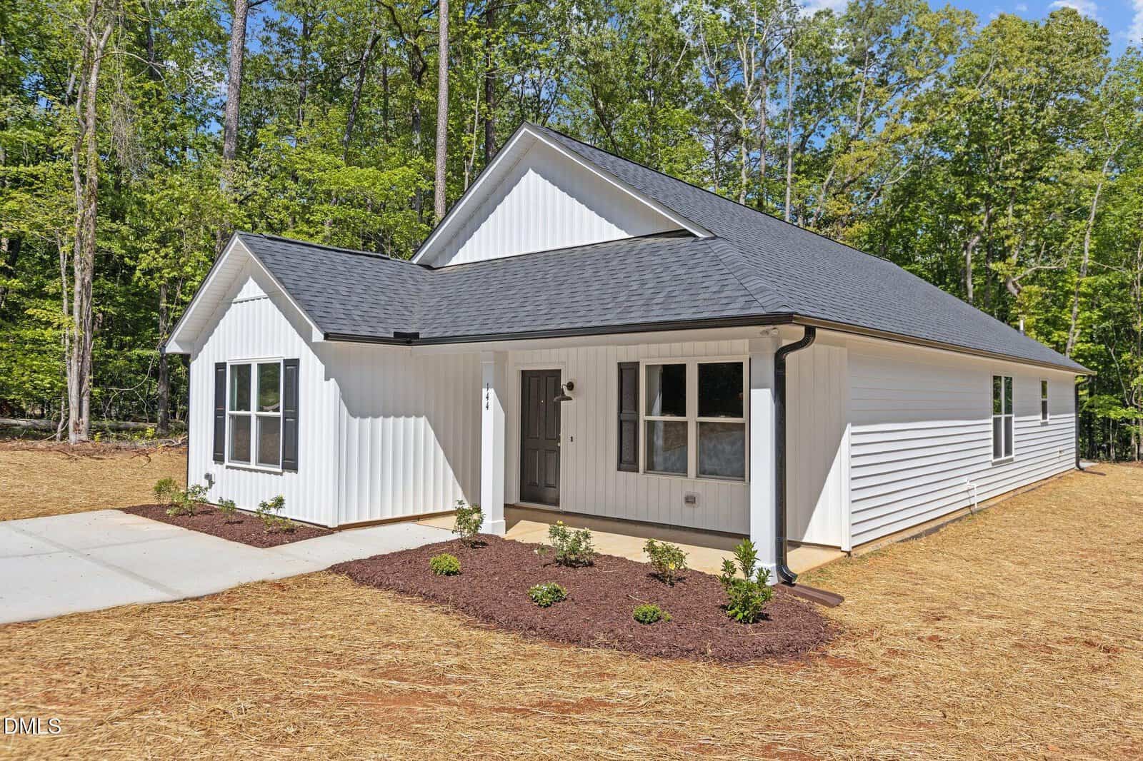 A modern single-story white house with dark roof shingles, surrounded by newly planted shrubs and fresh mulch, sits on a lot with dry straw and tall green trees in the background under a blue sky.