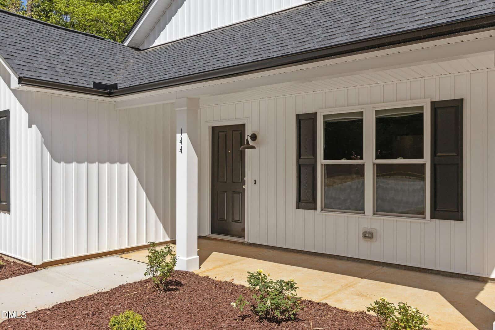 A modern white house exterior with vertical siding, black door, two windows with black shutters, a covered front porch, and small shrubs planted along a mulched garden bed.