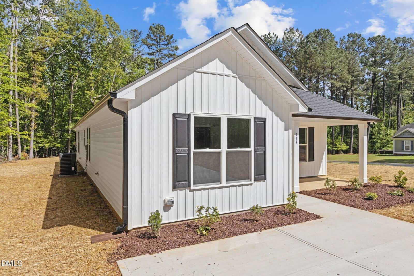 A modern single-story white house with black window shutters, a small front porch, and a neatly landscaped yard, surrounded by trees under a partly cloudy sky.