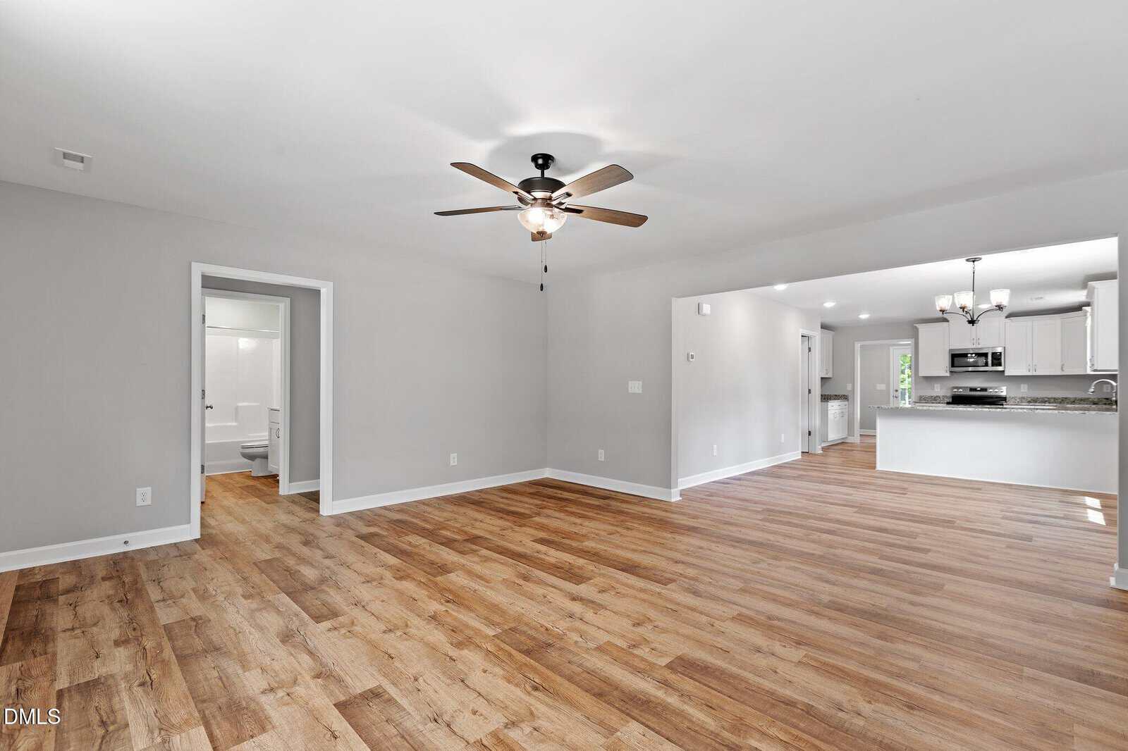 Bright, open living area with light wood flooring, neutral gray walls, ceiling fan, and recessed lighting. The space flows into a modern kitchen with white cabinets and a dining area; a bathroom is visible through a doorway.