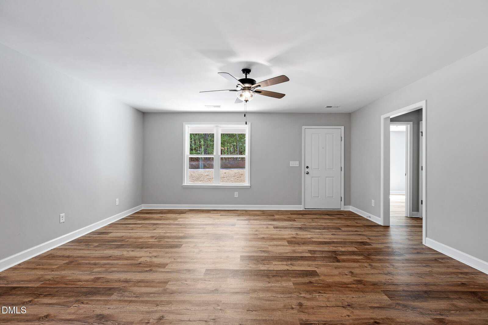 A spacious, empty living room with light gray walls, wood-style flooring, a ceiling fan with lights, a window, and a white door. An open doorway leads to another room on the right.