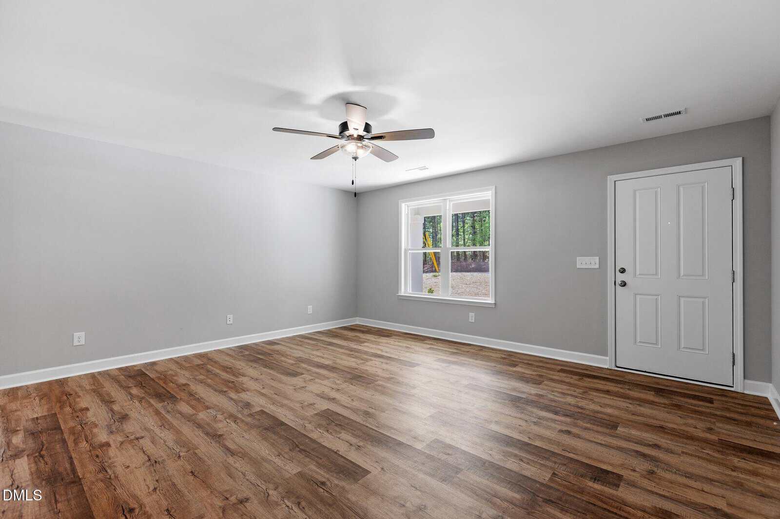 A bright, empty room with light gray walls, wood-look flooring, a ceiling fan with lights, a white door, two windows, and a view of greenery outside.
