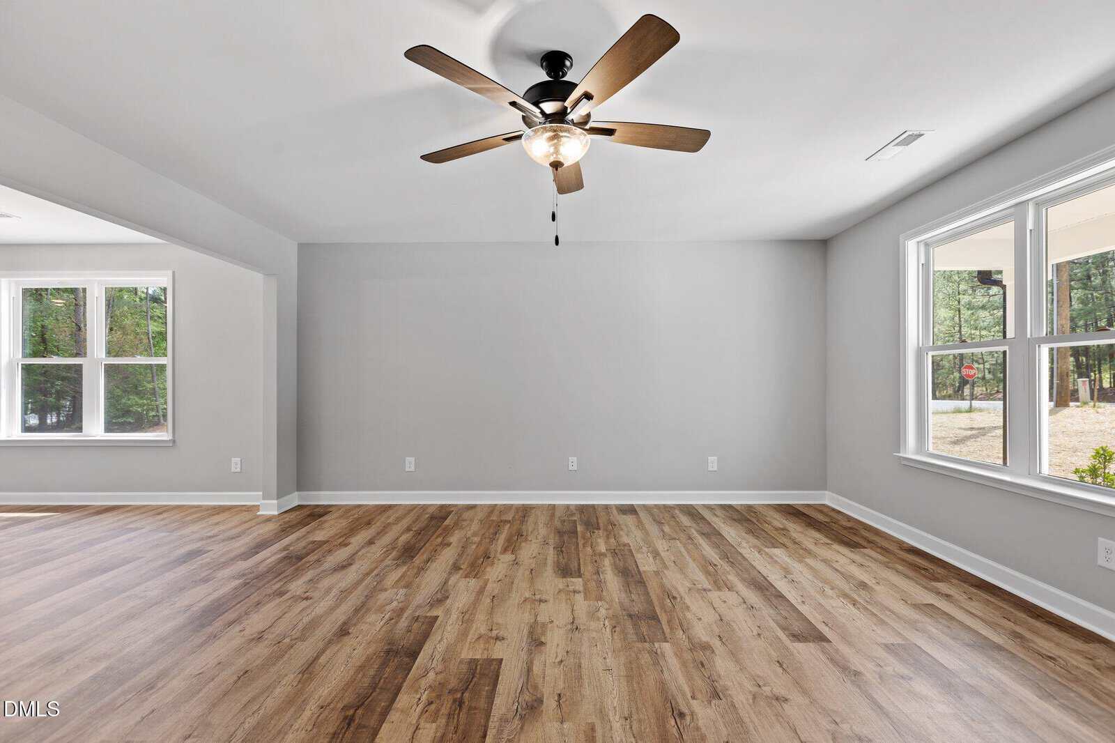 A spacious, empty room with light gray walls, wood-style flooring, a ceiling fan with lights, and large windows letting in natural light.