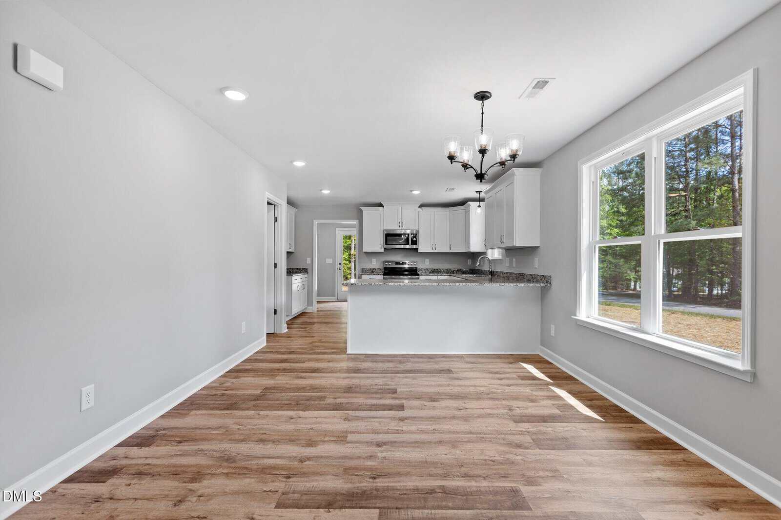 Bright, modern kitchen and dining area with light gray walls, wood-style flooring, white cabinetry, stainless steel appliances, a dark chandelier, and large windows overlooking a green outdoor space.