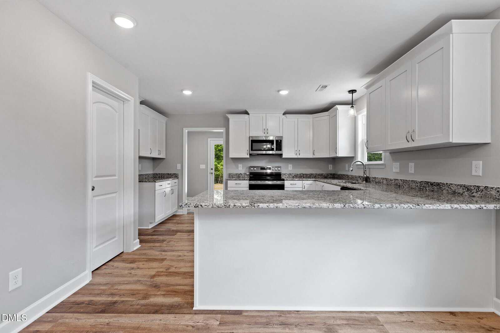 Modern kitchen with white cabinets, granite countertops, stainless steel appliances, wood flooring, and recessed lighting. A window above the sink and an open doorway lead to another room.