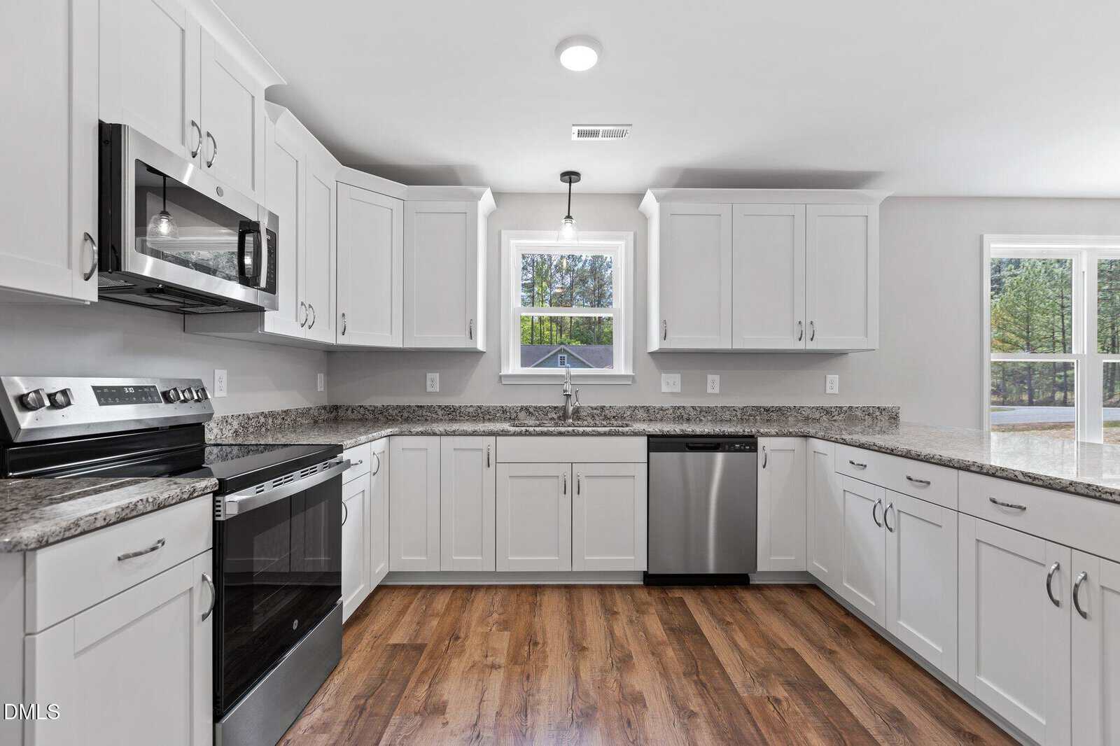 Modern kitchen with white cabinets, stainless steel appliances, granite countertops, and wood flooring. A window above the sink lets in natural light, and there are large windows to the right with a view outside.