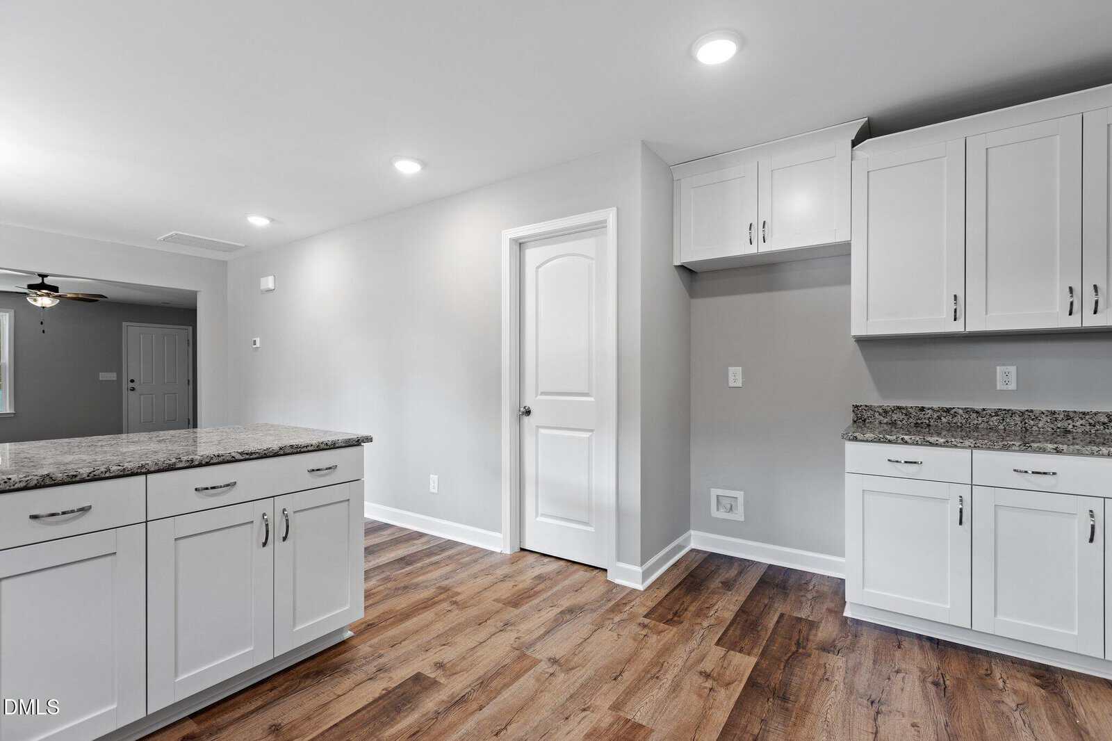 Modern kitchen with white cabinets, granite countertops, wood flooring, and recessed lighting. A white door is centered, while countertops and cabinets line the walls. The space appears clean and spacious.