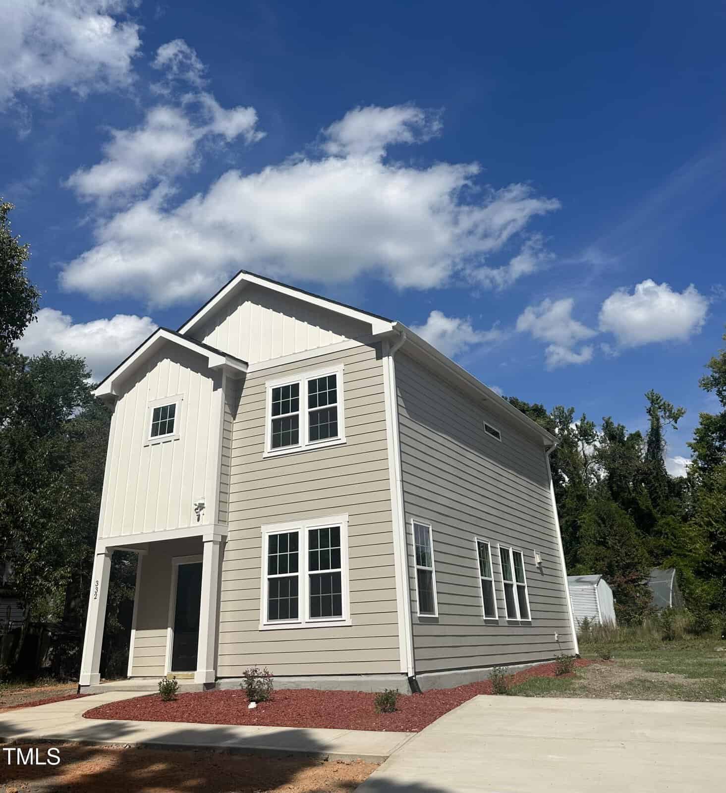 A modern two-story beige house with white trim and multiple windows sits under a partly cloudy blue sky. The front yard features red mulch landscaping and a concrete driveway, surrounded by trees and greenery.