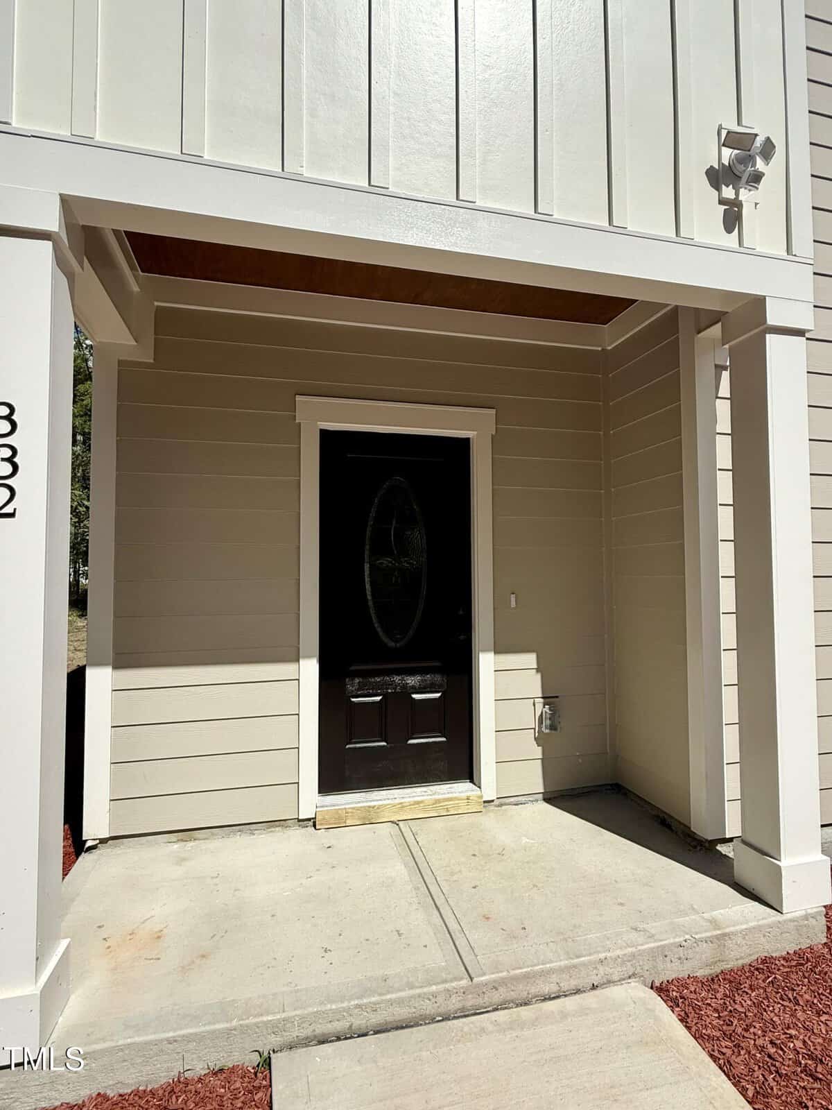 A covered front entryway of a beige house with vertical and horizontal siding, a black door with an oval glass window, concrete walkway, red mulch, and a security camera mounted on the wall.