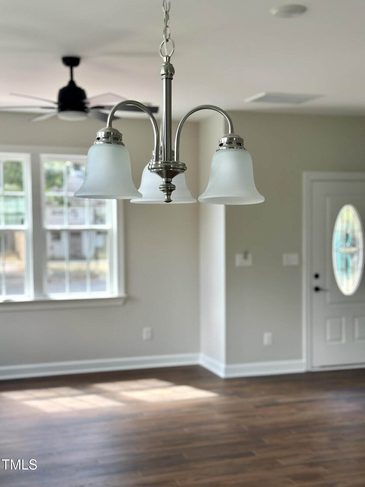 A brushed nickel chandelier with three frosted glass shades hangs in a bright room with hardwood floors, white walls, a window, and a white front door with an oval window.