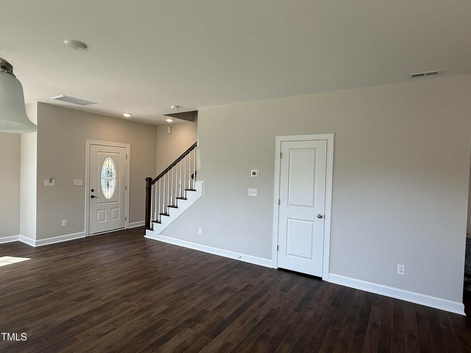 A spacious room with dark wood floors, light beige walls, a white front door with an oval window, and a white door to the right. A staircase with a dark railing is near the front door.