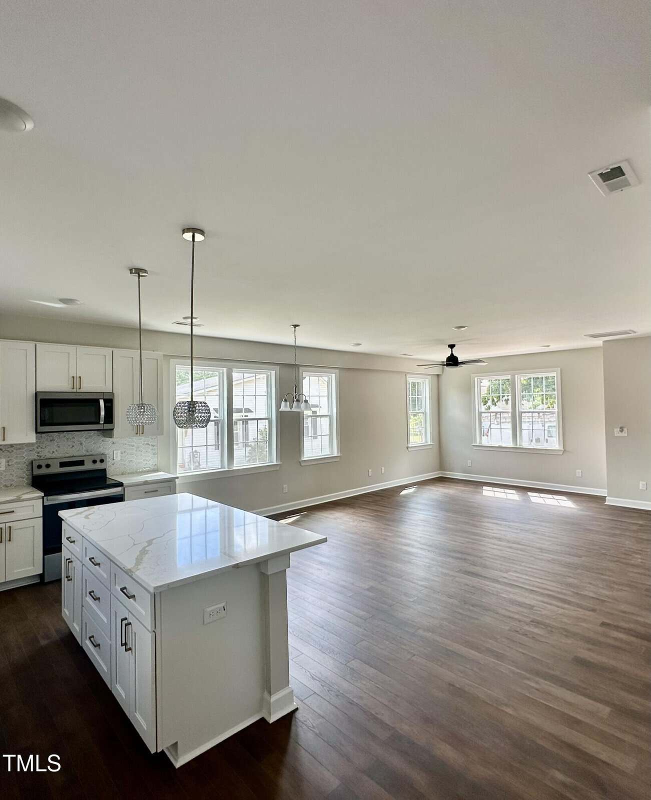 Bright, open-concept kitchen and living space featuring hardwood floors, large windows, white cabinets, stainless steel appliances, pendant lights over a marble island, and neutral walls.
