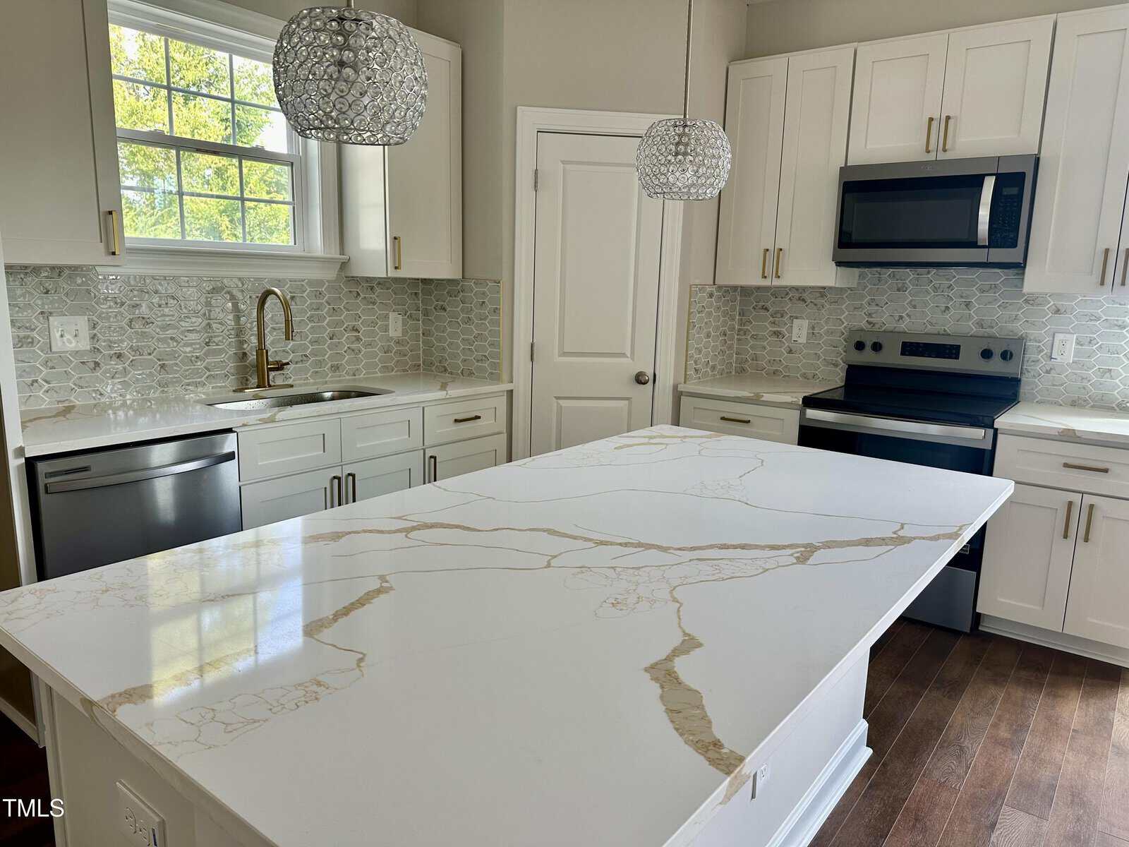 Modern kitchen with white cabinets, stainless steel appliances, mosaic tile backsplash, gold faucet, and large white marble island with tan veins under two decorative pendant lights. Wood flooring and window with greenery outside.