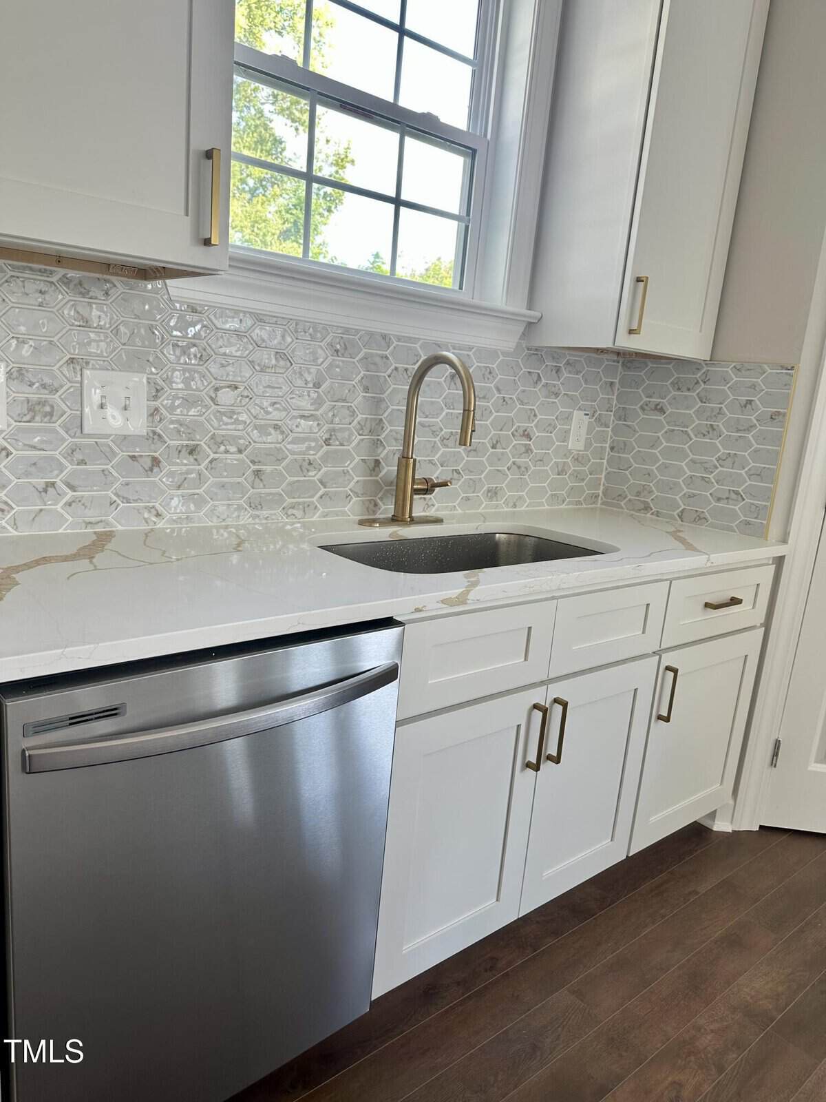 Modern kitchen with white cabinets, gold hardware, a stainless steel dishwasher, marble countertop, under-mount sink with a gold faucet, and a geometric tile backsplash. Sunlight streams in through a window above the sink.