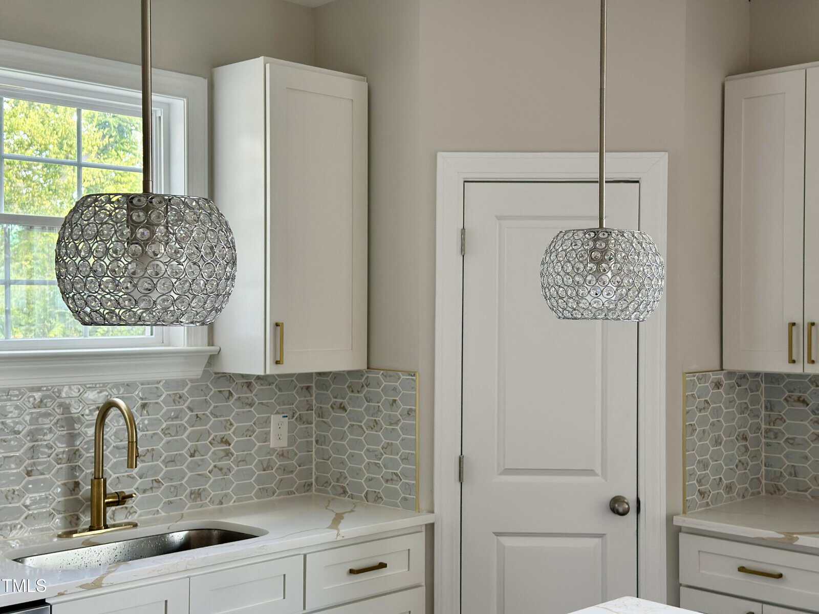 Modern kitchen with white cabinets, brass hardware, a gold faucet, geometric gray backsplash tiles, and two decorative pendant lights with circular crystal patterns hanging above a white countertop.