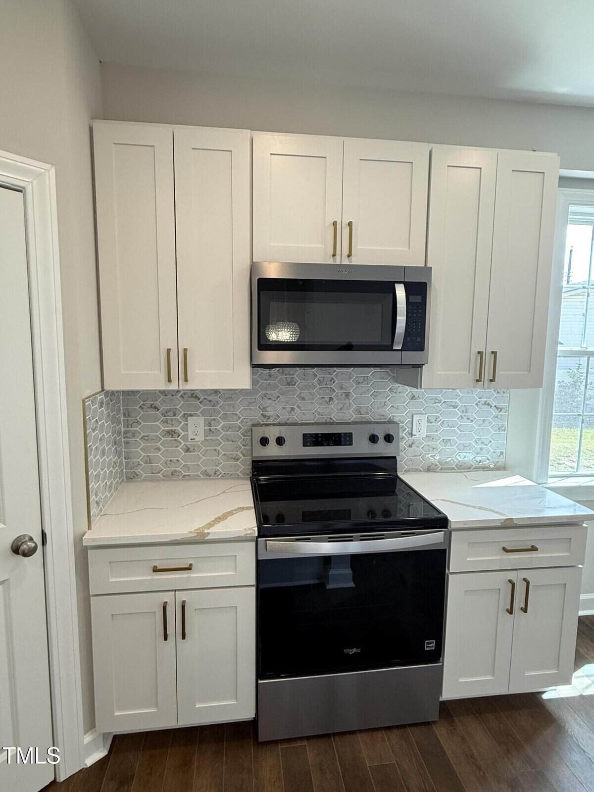 A modern kitchen with white cabinets, gold handles, a black electric stove, and a black microwave. The backsplash has a geometric tile pattern, and there is a window to the right letting in natural light.