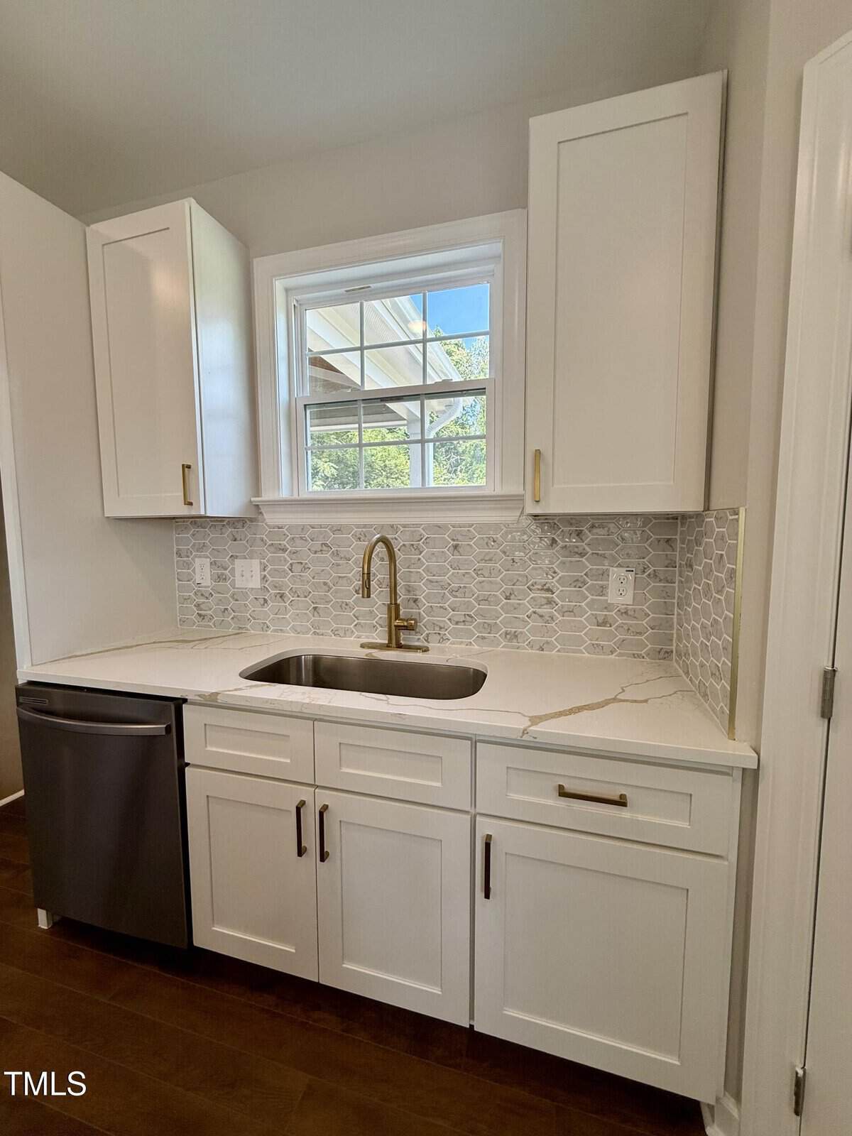 Modern kitchen with white cabinets, gold faucet, stainless steel dishwasher, and a gray hexagonal tile backsplash. A single window above the sink lets in natural light. White marble countertop with subtle veining.