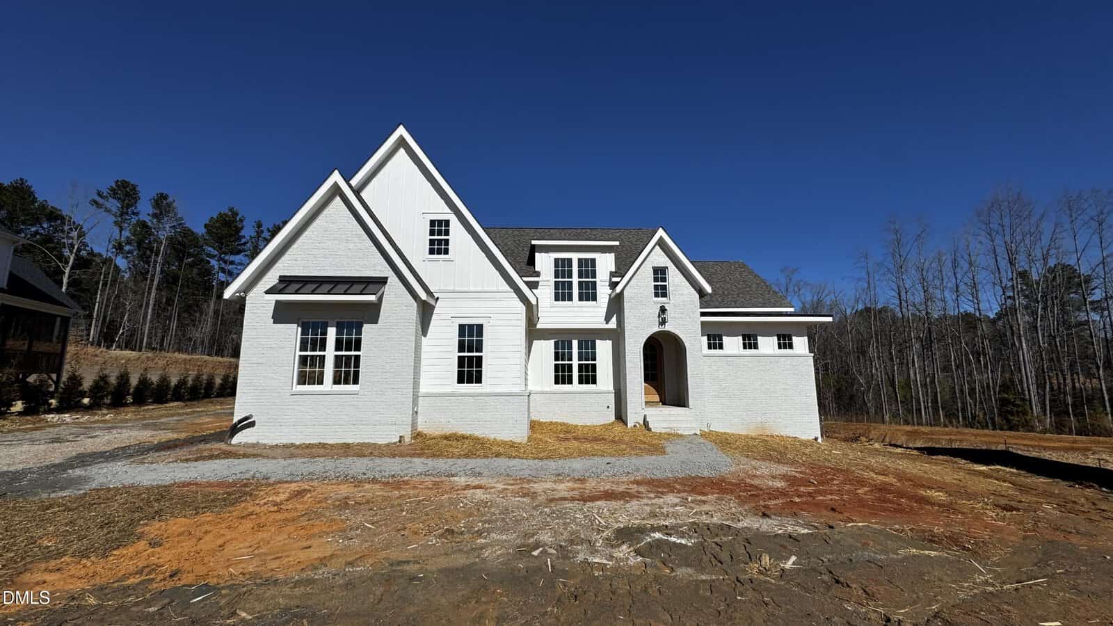 A newly constructed white brick house with multiple gables and arched entryway sits on a bare, reddish-brown dirt lot under a clear blue sky, surrounded by leafless trees and sparse landscaping.
