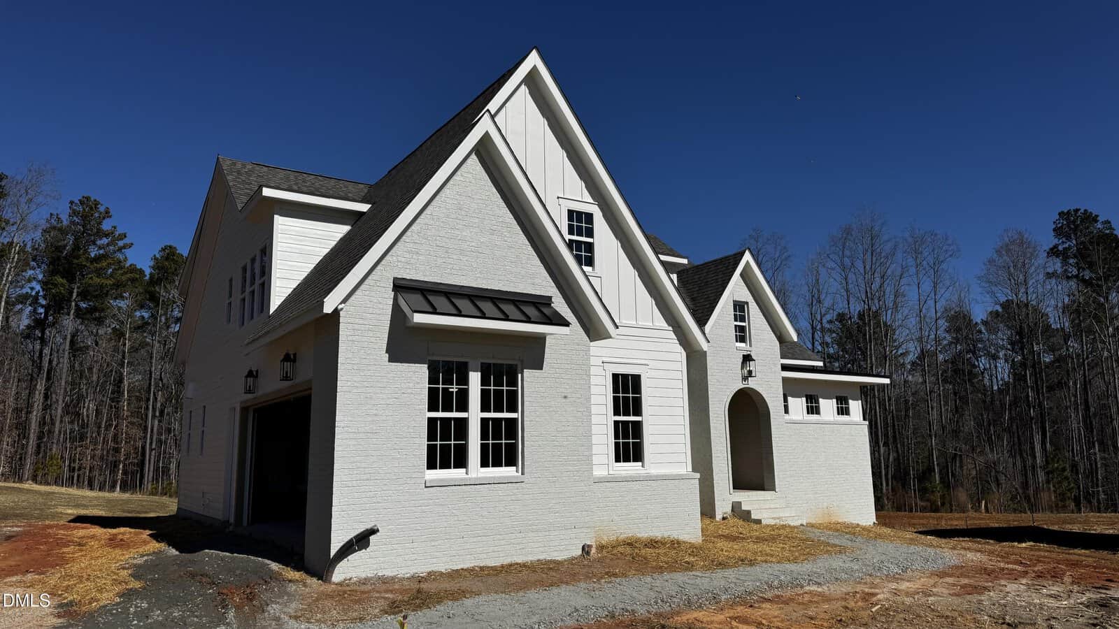 A newly constructed white brick house with steep gabled roofs, multiple windows, and an arched entryway, set on a bare lot with some trees in the background under a clear blue sky.