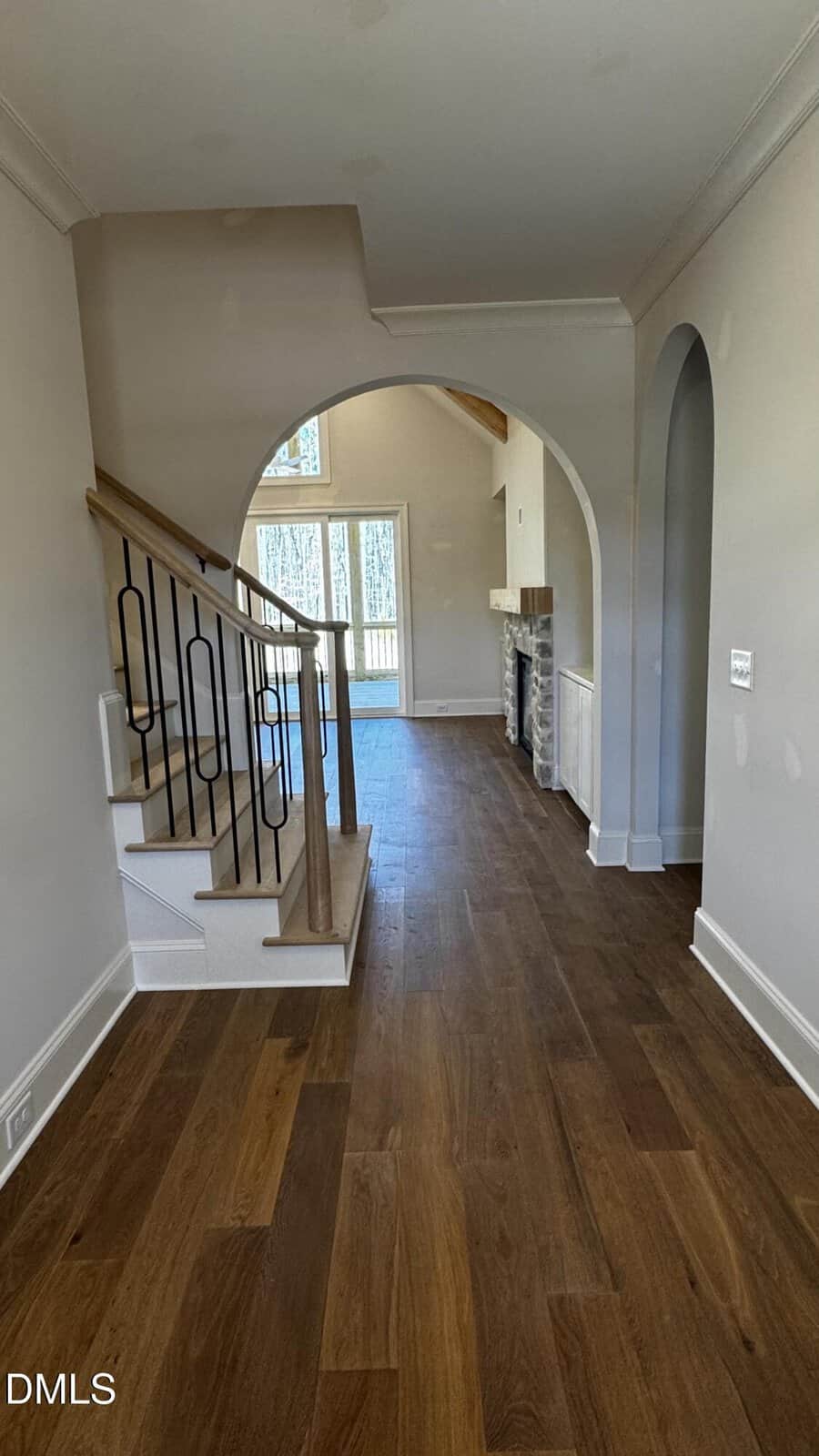View of a modern home interior with wooden floors, a staircase with black railings on the left, an arched doorway, and a living area with large windows and a fireplace in the background.