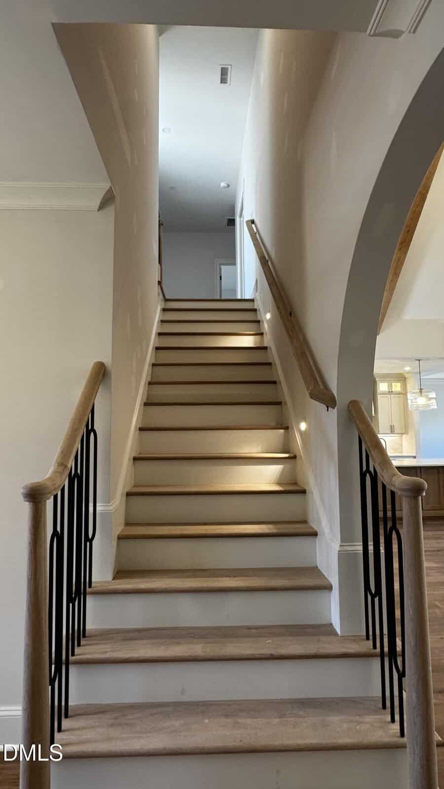A straight staircase with wooden handrails and black balusters leads up to a landing in a well-lit, modern home. Walls are painted light, and patchwork is visible. An arched entryway is on the right.