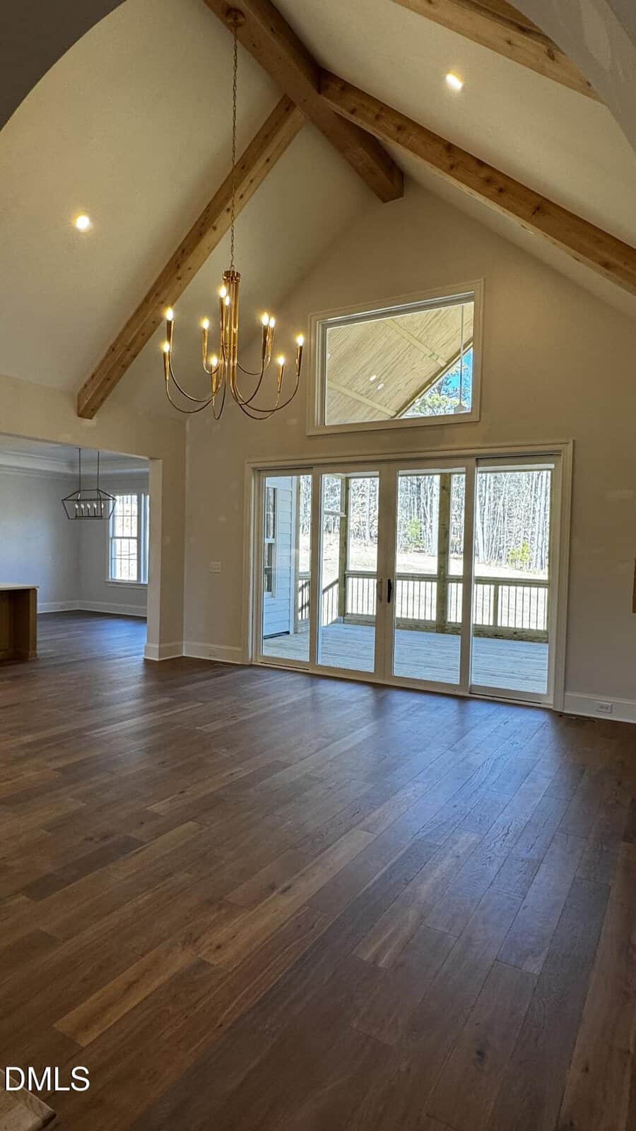 Spacious living area with vaulted ceiling, exposed wooden beams, large window above glass doors, modern chandelier, hardwood floors, and view into adjacent dining space with pendant light. Outdoor deck visible through the doors.