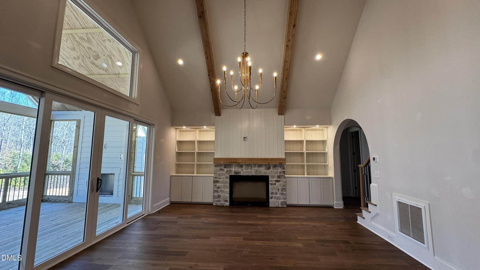 A bright living room with high ceilings, wooden beams, a modern chandelier, built-in shelves, a stone fireplace, and large windows opening to a covered deck.