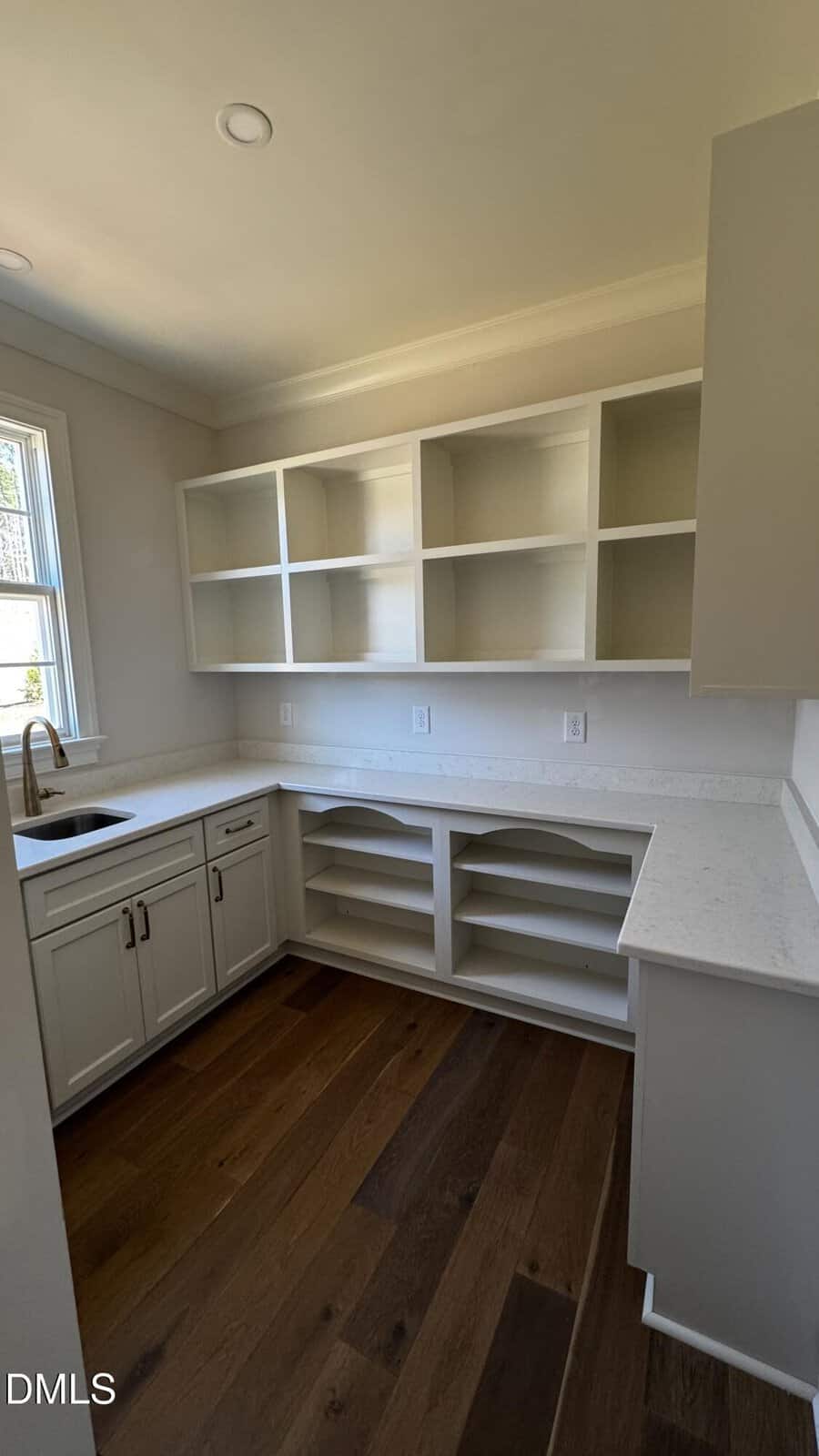 A bright, modern pantry or laundry room with white cabinets, open shelves, a sink under a window, light countertops, and dark wood flooring. Natural light streams in from the window.