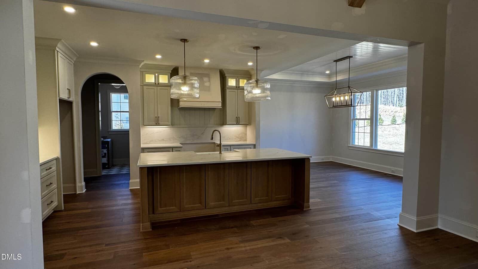 A modern kitchen with a wooden island, marble countertops, pendant lights, white cabinets, and an adjacent dining area with large windows letting in natural light. The floor is dark wood.