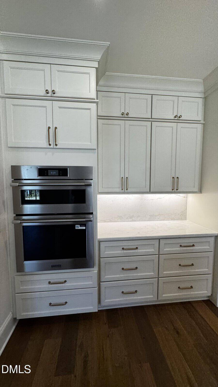 Built-in stainless steel double oven next to white cabinets and drawers with gold handles, under-cabinet lighting, and a white marble backsplash, all set on a dark hardwood floor.