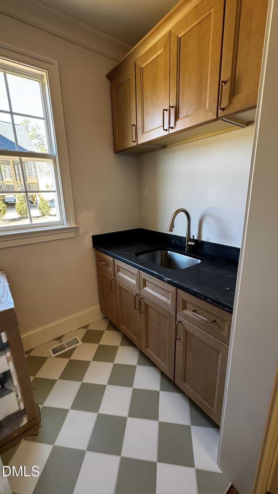 Small laundry or utility room with light wood cabinets, black countertop, stainless steel sink, chrome faucet, checkerboard tile floor, and a window letting in natural light.