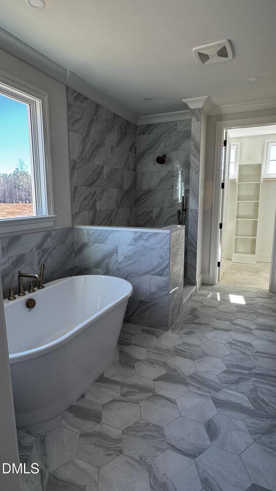 Modern bathroom with a white freestanding tub, hexagonal floor tiles, marble-look wall tiles, and a walk-in shower. Sunlight streams through a window and an open door leads to a walk-in closet.