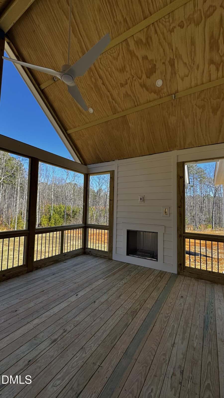 A covered wooden porch with a ceiling fan, built-in fireplace, and open views of a grassy yard and trees under a clear blue sky.